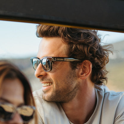 Man wearing Mireye Roti sunglasses in deep forest, side profile in an open-top car showing the bold rectangular frame.
